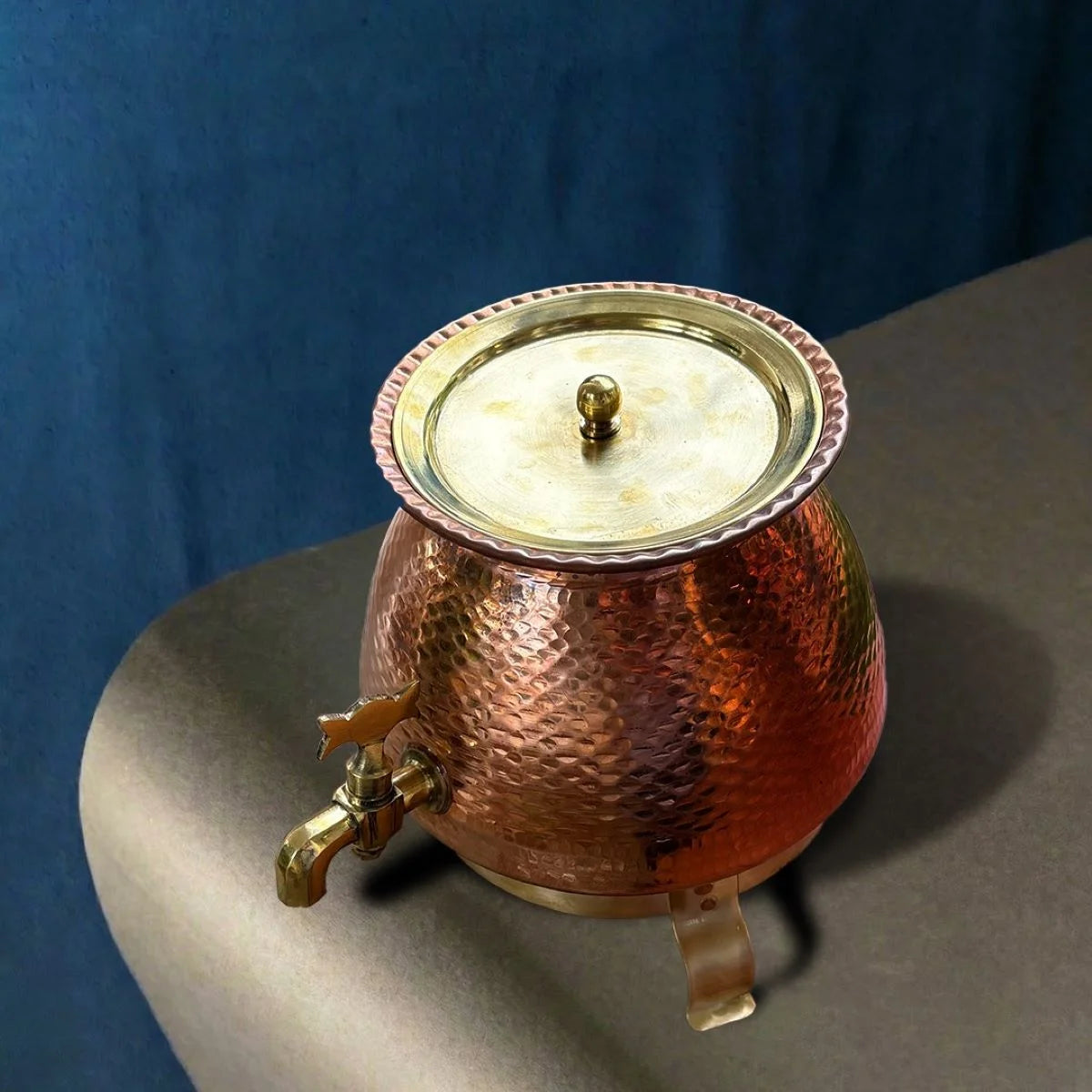 Copper water container with a lid, tap and a stand, on a gray surface against a blue background. Zishta Traditional Serveware