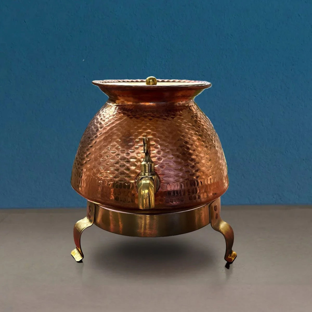 Copper water pot with a spout on a gray surface and blue background. Zishta Traditional Serveware