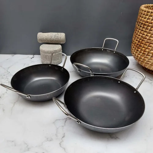 Three black pure iron flat bottom kadai with handles on a marble surface with a gray wall and woven basket in the background-Zishta Traditional Cookware