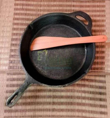 Black cast iron skillet with a wooden spoon on a woven mat-Zishta Traditional Cookware