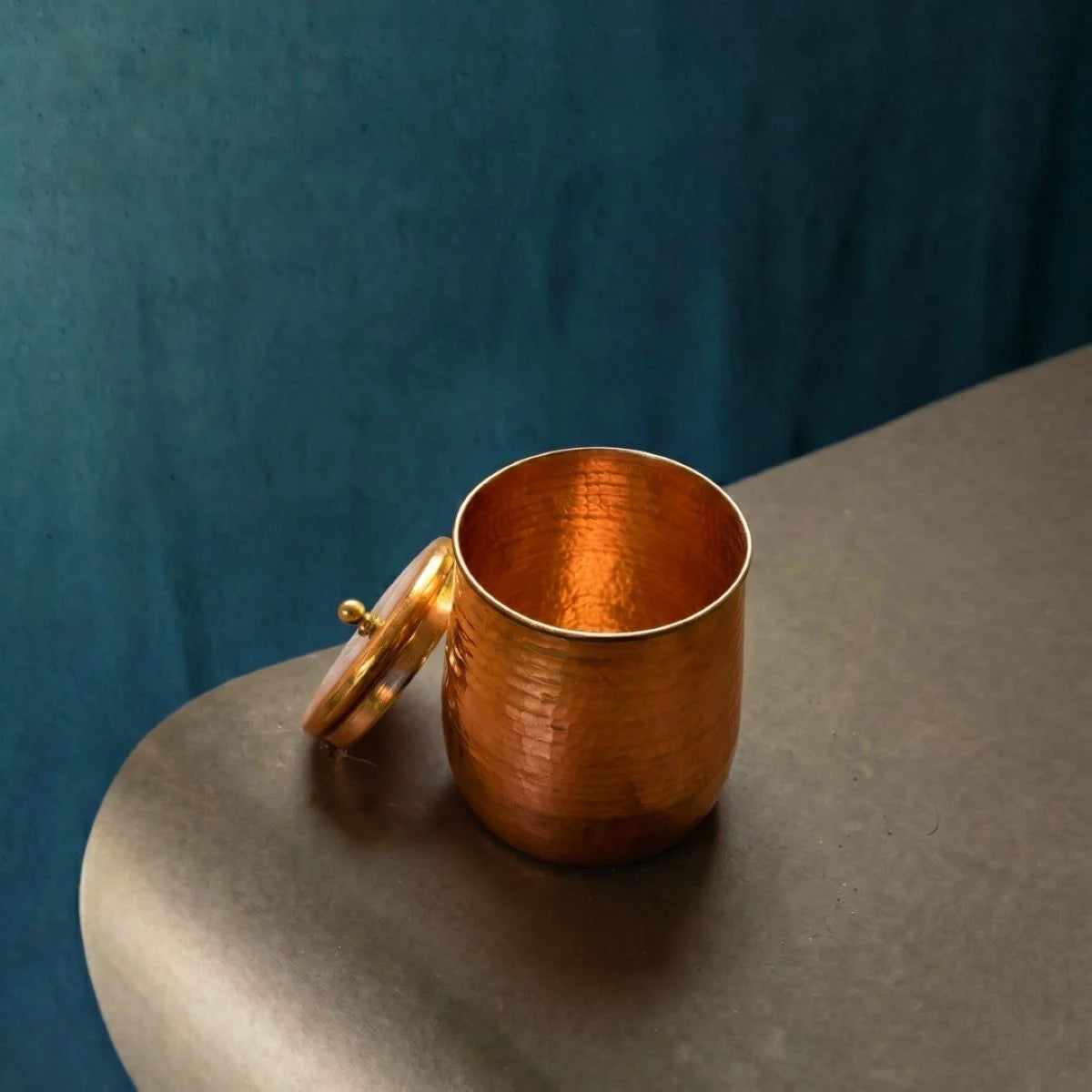 Copper cup with lid on a gray surface against a blue background. ZIshta Traditional Drinkware.