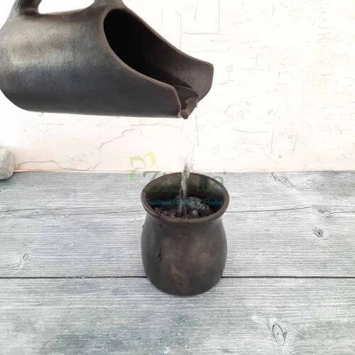 Manipur Black Pottery Jug pouring water into a Manipur Black Pottery Mug on a wooden surface-Zishta Traditional Serveware